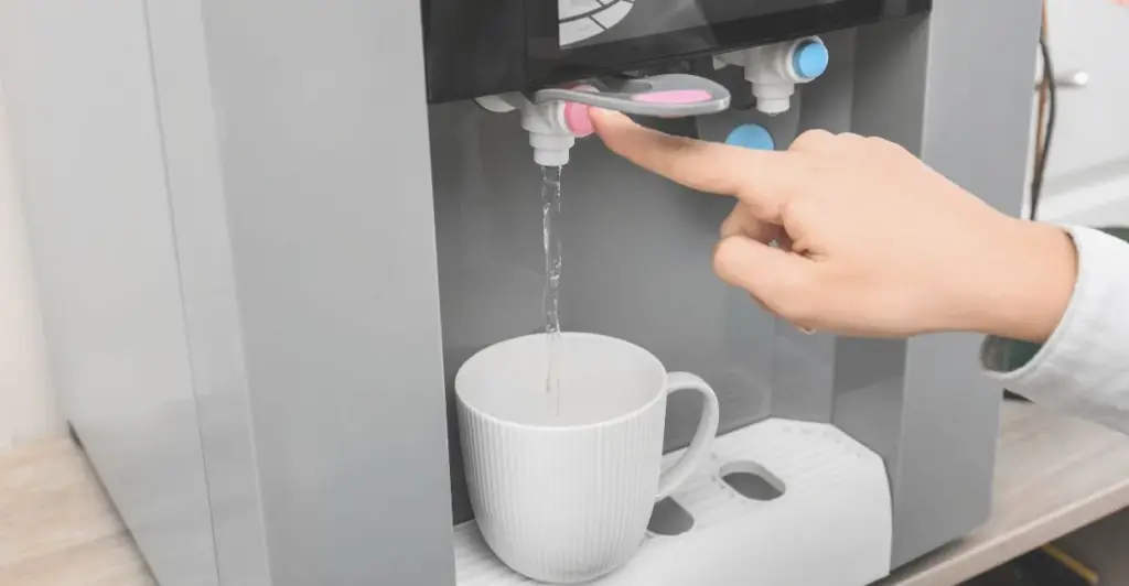 Filtro de água branco instalado em pia de cozinha, com mão pressionando o botão para encher um copo transparente com água purificada. Imagem do CANVA