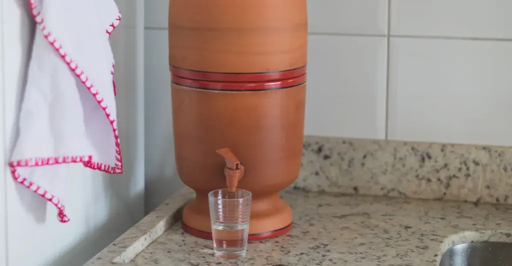 Filtro de barro tradicional derramando água cristalina em um copo, demonstrando o processo natural de filtragem.