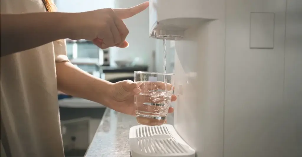 Pessoa enchendo um copo de água em um purificador de água moderno, visto de lado, mostrando apenas os braços, o copo transparente e o filtro branco. quais os melhores purificadores de agua