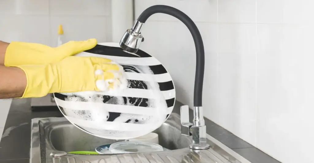 Braços de pessoa lavando louça em pia com torneira elétrica Hydra Patinho, jorro de água quente saindo do dispositivo, pratos e talheres sendo higienizados com eficiência.